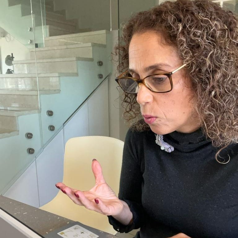 אישה עם שיער מתולתל ומשקפיים מביטה בידה כשהיא יושבת ליד שולחן בחדר מודרני, כשברקע נראים מדרגות זכוכית.