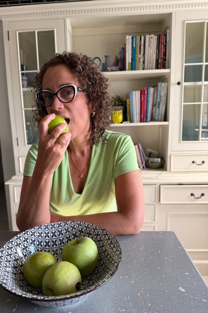 אישה עם שיער מתולתל ומשקפיים אוכלת תפוח ירוק על דלפק המטבח, כשקערית תפוחים מונחת לפניה ומדף ספרים ברקע.